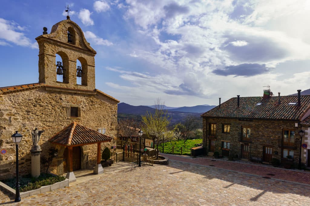 Imagen de la iglesia de San Miguel Arcángel y una casa típica de piedra en La Hiruela, Madrid, rodeadas de vegetación